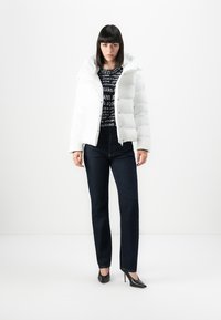 Vrouw in een witte pufferjack, met een zwart patroontrui, donkere jeans en zwarte hoge hakken, staande tegen een witte achtergrond.