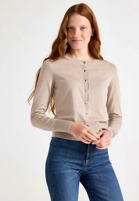 Béžový pletený kardigan s kulatým výstřihem, zapínáním na knoflíky vpředu a žebrovanými manžetami. Kombinovaný s modrými džínami, měkká textura a úzký střih.