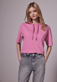 Růžový top s krátkým rukávem, kapucí a šňůrkami, spárovaný s šedými džíny s vysokým pasem, které mají vybledlou texturu.