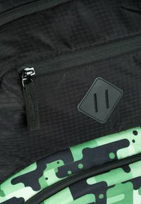 Tessuto nero testurizzato con un fondo verde a motivo mimetico. Presenta una tasca con zip e un'etichetta di gomma rettangolare con due linee.