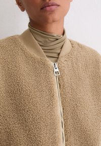 Fleece-Bomberjacke in Beige mit strukturierter Außenseite. Verfügt über einen Reißverschluss und eine weiche, glatte Innenschicht in einem helleren Farbton.