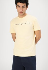 Tricou galben din bumbac, cu mâneci scurte, având logo-ul "TOMMY JEANS" în negru pe piept, asortat cu pantaloni scurți închisi la culoare.