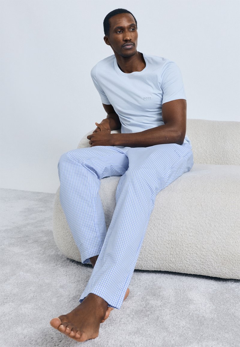 Vaaleansininen puuvillainen t-paita kirjotulla logolla, yhdistetty sinisiin ja valkoisiin ruudullisiin pyjamahousuihin, istuen teksturoidulla, neutraalinvärisellä pinnalla.