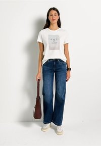 T-shirt blanche avec un imprimé graphique léger, jeans larges bleus et baskets beiges. Le modèle tient un long sac à main marron.