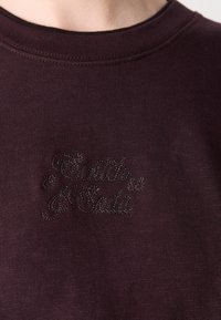 Sudadera burdeos con cuello de canalé y logotipo "Bati" bordado. Fabricada en un tejido suave con una textura lisa, cuenta con un diseño de cuello redondo.