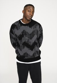 Grau und schwarz gemusterter Strickpullover mit Rundhalsausschnitt, der Zickzack- und vertikale Linienmuster aufweist. Über einem weißen Hemd getragen. Schwarze Hose.