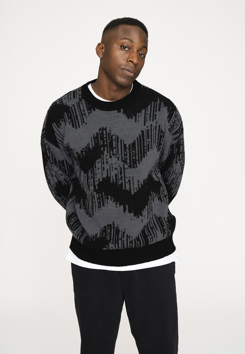 Grau und schwarz gemusterter Strickpullover mit Rundhalsausschnitt, der Zickzack- und vertikale Linienmuster aufweist. Über einem weißen Hemd getragen. Schwarze Hose.