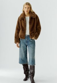Bruine nep bontjas met een brede kraag, gecombineerd met lichtblauwe, cropped jeans en bruine kniehoge laarzen. Eenvoudig wit t-shirt eronder.