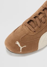 Brązowe zamszowe trampki z teksturowaną powierzchnią, z białą gumową podeszwą i haftowanym logo na boku. Sznurowadła są w pasującym odcieniu brązu.