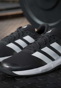Paire de chaussures de sport noires avec des rayures blanches et des lacets, placées sur un sol de gym en caoutchouc texturé près d'une plaque de poids.