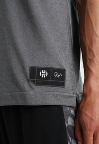 Chemise de sport grise à texture lisse, avec une étiquette noire arborant un logo et des détails signature. Comprend une fente latérale pour plus de confort.