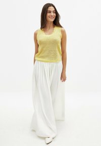 Suéter amarillo sin mangas con un diseño de punto texturizado, combinado con una falda larga blanca fluida. La modelo está de pie sobre un fondo neutro.