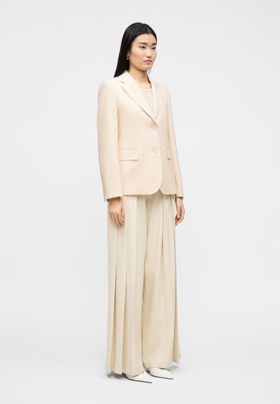 Γυναίκα με μακριά σκούρα μαλλιά που φοράει ένα κρεμ σακάκι, κρεμ φαρδιά παντελόνια με πιέτες και λευκά μυτερά παπούτσια σε απλό φωτεινό φόντο.