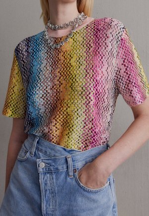 Femme portant un haut à manches courtes multicolore à motifs en zigzag, un jean bleu taille haute et des colliers de cristal superposés.
