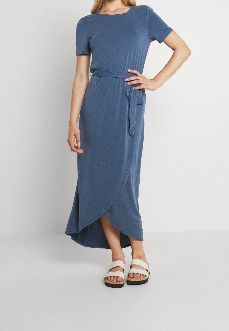 Robe longue bleu avec des manches courtes et une taille cintrée. Présente un design croisé et une fente sur le côté. Associée à des sandales légères.