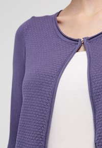 Cardigan violet texturé avec un col rond et une fermeture frontale. Porté sur un haut de couleur claire, mettant en valeur les couches contrastantes.