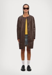 Brauner Lederhut mit einer gewebten Akzentierung auf der Vorderseite, kombiniert mit einem gelben Pullover, einem mehrfarbigen Fransenrock und schwarzen Schuhen.