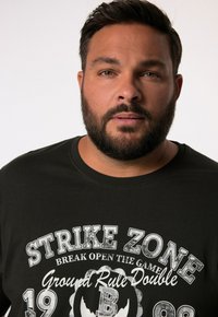 Tmavě zelené bavlněné tričko s kulatým výstřihem, se zaobleným bílým potiskem a grafikou zdůrazňujícími "STRIKE ZONE" a "Ground Rule Double."
