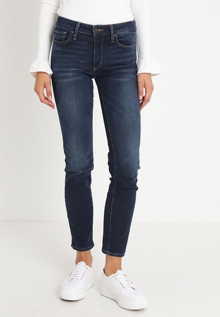 Femme portant un jean skinny bleu foncé, un haut blanc à manches longues avec des poignets volantés et des baskets à plateforme blanches, debout sur un sol blanc.