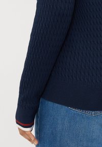 Tummansininen neulepaita, jossa on teksturoitu timanttimalli, punavalkoiset ribbauskäyttäjät ja kapea helma farkkujen päällä.