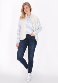 Fleece brezrokavnik v kremni barvi, položen čez modro-belo črtasto srajco z dolgimi rokavi; kombiniran s temnimi ožjimi jeans hlačami in belimi supergami.