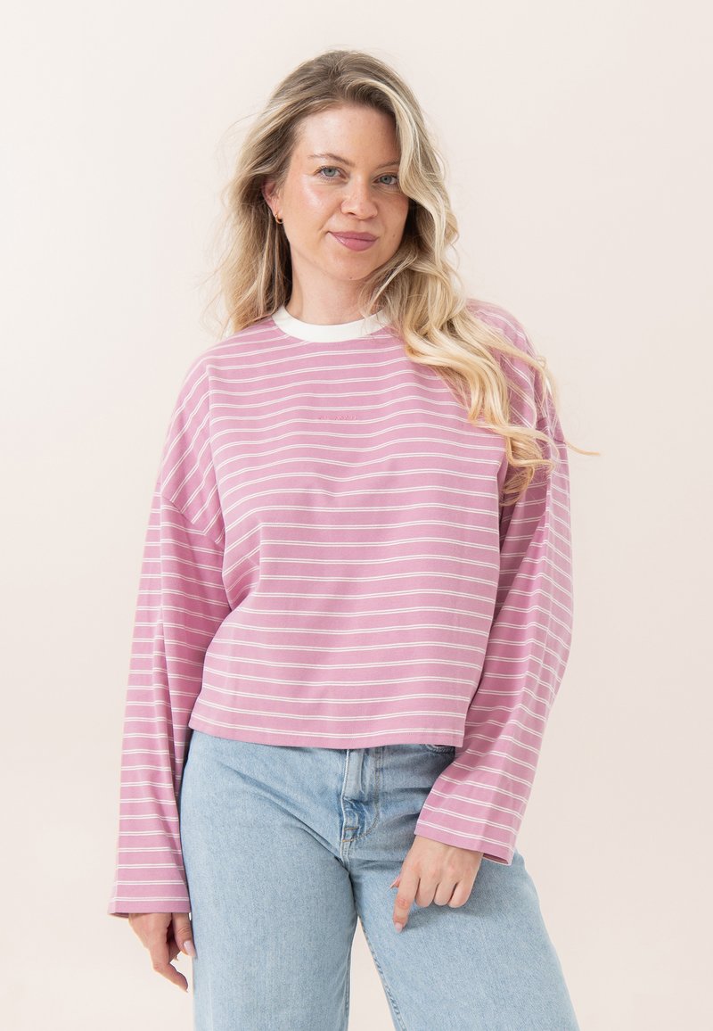 Ξανθιά γυναίκα με ανοιχτά μαλλιά που φοράει ροζ και λευκή ριγέ μπλούζα με μακριά μανίκια και ανοιχτόχρωμα μπλε τζιν, στέκεται μπροστά σε απλό φόντο.