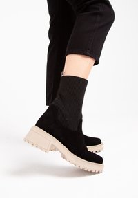 Czarne zamszowe botki za kostkę z ozdobnym prążkowanym mankietem w formie skarpety oraz jasnobeżowymi masywnymi podeszwami. Posiadają lekko podniesiony obcas i teksturowaną podeszwę.