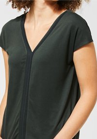 Top de color verde oscuro con escote en V y mangas cortas, que presenta un ribete texturizado en negro a lo largo del escote. Tejido suave, corte relajado.