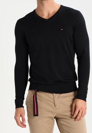 Mann trägt schwarzen V-Ausschnitt Pullover mit langen Ärmeln und kleinem Logo auf der Brust sowie beige Hose mit rot gestreiftem Gürtel, der herunterhängt.