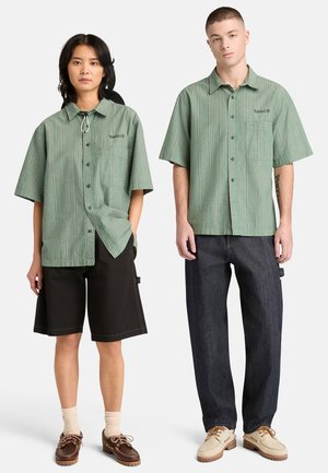 Kaksi mallia, jotka käyttävät vihreäraidallisia, lyhythihaisia nappipaitoja; yhdellä mustat shortsit ja ruskeat kengät, toisella tummat farkut ja beiget kengät.