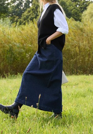 Donna che cammina in un campo erboso indossando camicia bianca, gilet nero, lunga gonna blu scuro, stivali neri, tenendo una borsa bianca.
