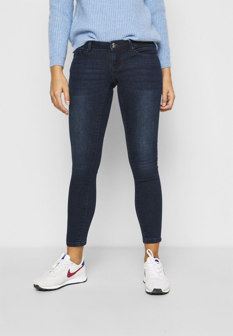 Donkerblauwe skinny jeans met een gladde textuur, voorzien van een sluiting met knoop aan de voorkant, ceintuurlussen en subtiele vervaging op de dijen.