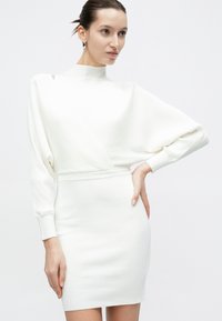 Frau trägt ein weißes, geripptes Strickkleid mit langen Fledermausärmeln und hohem Kragen und posiert mit einer Hand in der Hüfte.