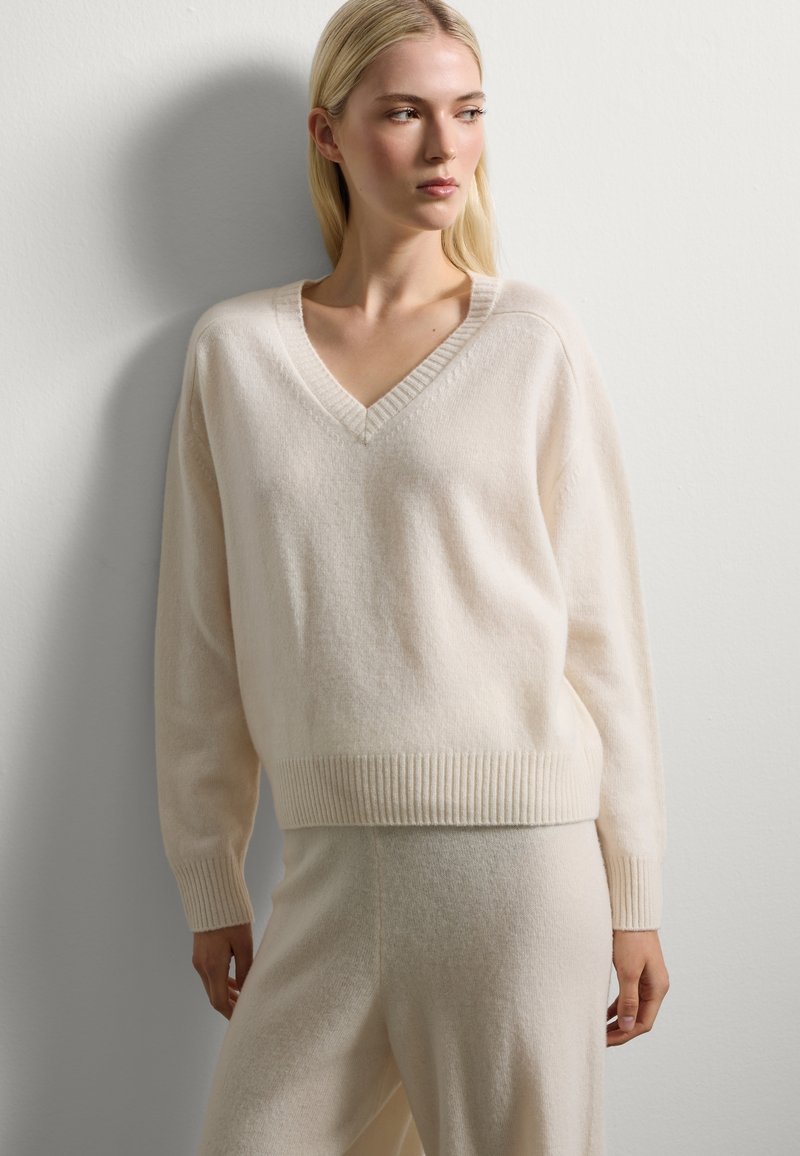 Maglione a V in morbido crema, realizzato in un tessuto leggero. Presenta polsini e fondo a coste, con una vestibilità rilassata e una trama liscia.