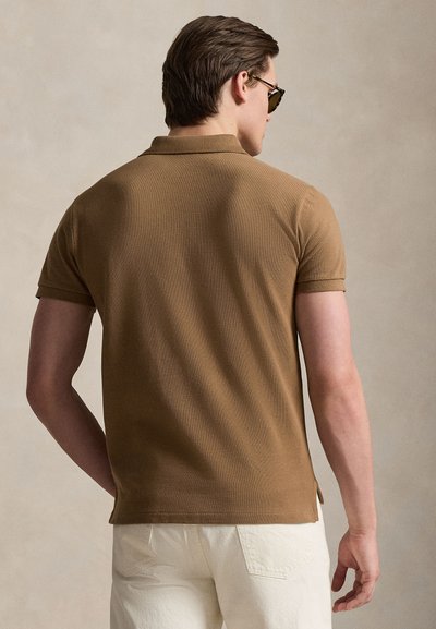 Καφέ κοντομάνικο πόλο πουκάμισο με υφασμάτινη υφή και διπλωμένο γιακά, συνδυασμένο με ανοιχτόχρωμο παντελόνι. Διακριτικές πλευρικές σχισμές στο τελείωμα.