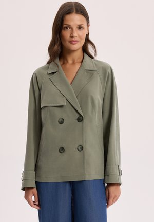 Kobieta w zielonkawo-szarej, dwurzędowej marynarce z detalami guzików oraz w niebieskich spodniach o szerokich nogawkach, stojąca na jednolitym jasnym tle.