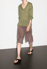 Groene gebreide v-hals trui, bruine suède shorts en zwarte sandalen. De trui heeft een ruime pasvorm; de shorts zijn knielang met een zachte textuur.