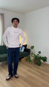 Man with curly hair stands in living room wearing white long-sleeve shirt, dark jeans, and brown loafers near green sofa and indoor plants.