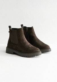 Donkerbruine suède enkelboots met zwarte chunky zolen, zij-elastische panels en binnenritsen op een witte achtergrond.