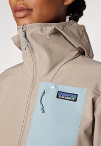 Lähellä kuva henkilöstä, joka on pukeutunut beigeen ja vaaleansiniseen Patagonia-takkiin, jossa on korkea kaulus ja näkyvä vetoketjuhäntä.