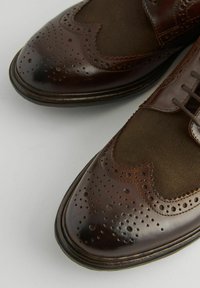 Braune Leder- und Segeltuch-Schnürschuhe im Wingtip-Stil haben eine spitze Zehenform, durchbrochene Details und eine strukturierte Oberfläche. Das Design umfasst Schnürsenkel und kontrastierende Materialien.