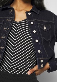 Veste en denim foncé avec des coutures claires, cinq boutons et une poche poitrine, portée par-dessus une chemise rayée noir et blanc à col en V.