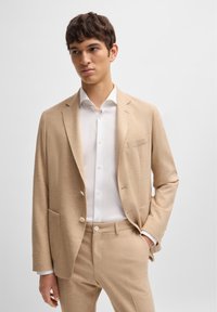 Veste de costume beige en tissu texturé, dotée d'un design à un bouton, de deux poches avant et d'un col cranté, assortie d'une chemise blanche.