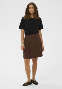 Femme aux cheveux bouclés portant un t-shirt noir "Less is more", une jupe marron au genou, des chaussures tissées noires et un bracelet en argent, se tenant sur fond blanc.