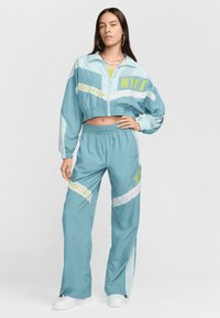 Trening din material albastru deschis Nike, cu jachetă scurtă și pantaloni lejeri. Logo-uri "Sportswear" și "Nike" în galben neon. Textură netedă.