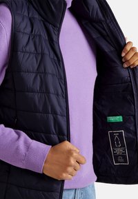 Osoba v fialovém svetr a modrých džínách, držící otevřenou černou prošívanou vestu, v níž je vidět zelenou nálepku "United Colors of Benetton".