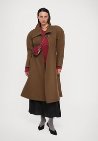 Cappotto di lana marrone con colletto alto e bottoni doppi, indossato su un top texture rosso e una gonna nera. Gli accessori includono una borsa a tracolla rossa.