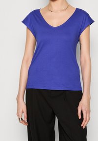 Person som bär en kortärmad, V-ringad lila skjorta, svarta byxor, ett guldhalsband och ringar på båda händerna, stående mot en enfärgad bakgrund.