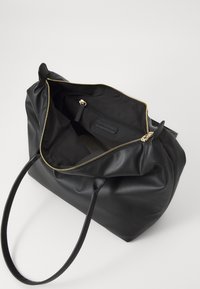 Sac fourre-tout en cuir noir avec une fermeture éclair dorée, texture douce et longues poignées. L'intérieur comprend une doublure en tissu noir avec une poche zippée.