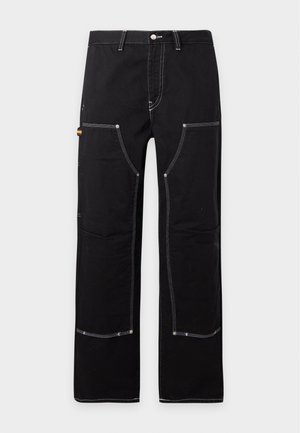 Homme debout les mains dans les poches, portant une veste et un pantalon noirs avec des coutures blanches et des baskets noires avec des lacets rouges sur fond blanc.